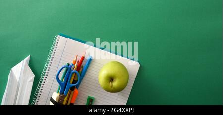 Open spiral notebook school tools on top with  apple and paper plane on green background with space for text. Top view. Panoramic composition. Stock Photo