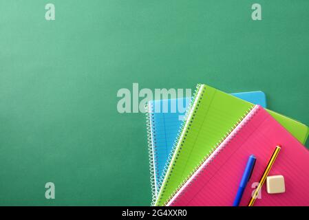 Three colored spiral notebooks with pencil and eraser pen on top on green background with space for text. Top view. Stock Photo