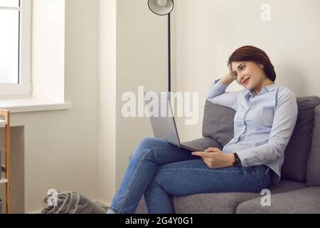 Smiling millennial woman looking at laptop screen relax on couch at home Stock Photo