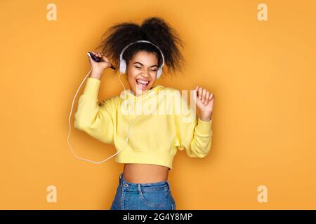Cheerful girl uses headphones and smartphone, listens music, sings song aloud on yellow background Stock Photo