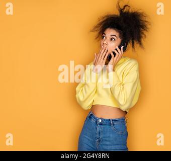 Girl is talking on the phone and looking shocked. Photo of african american girl on yellow background Stock Photo