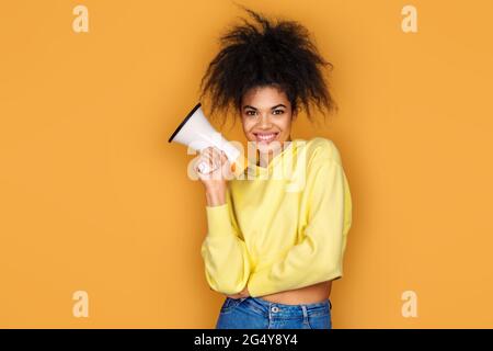 Smiling girl holds megaphone in hand. Photo of african american girl on yellow background Stock Photo