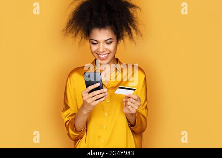 Young girl makes a payment, using a credit card and smartphone. Photo of african american girl on yellow background Stock Photo