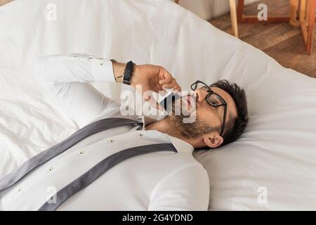 Businessman in eyeglasses and shirt talking on smartphone while lying on bed Stock Photo
