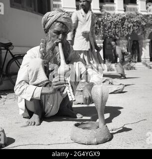1950s, historical, a snake charmer, with a python snake around his neck, squatting on the floor playing a musical instrument, a pungi, to another snake, a cobra, whose head emerges from a small basket,  India. Stock Photo