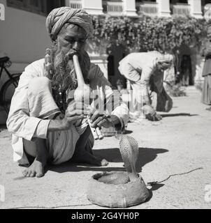 1950s, historical, a snake charmer, with a python snake around his neck, squatting on the floor playing a musical instrument, a pungi, to another snake, a cobra, whose head emerges from a small basket,  India. Stock Photo