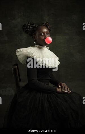 Portrait of medieval African young woman in black vintage dress with big white collar posing isolated on dark green background. Stock Photo