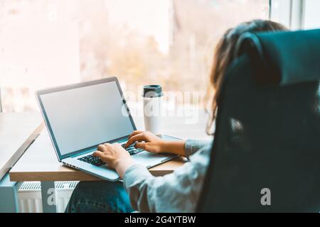 Woman working with a laptop at the modern bright office with a eco-friendly coffee cup, sitting at the desk. Blank white screen for text copy Stock Photo