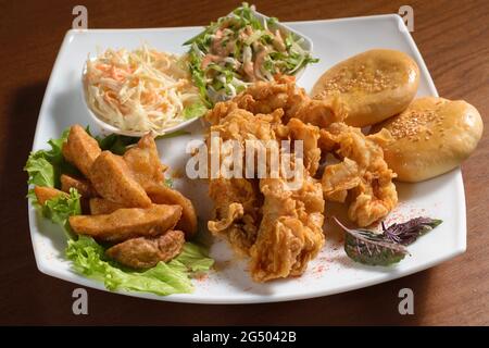 Crispy fried chicken pieces in breadcrumbs on a plate with potatoes, bread and salad, on a wooden table Stock Photo