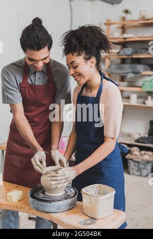 positive young african american couple in aprons shaping wet clay pot on wheel with hands in pottery Stock Photo