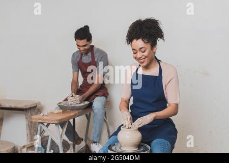 smiling young african american couple in aprons working with wet clay pots in pottery Stock Photo