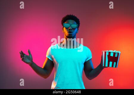 Image of young african man with the box of popcorn in his hands on bright pink neon background. Stock Photo