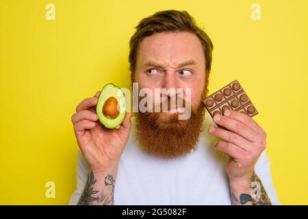 Doubter man with beard and tattoos holds avocado and chocolate Stock Photo