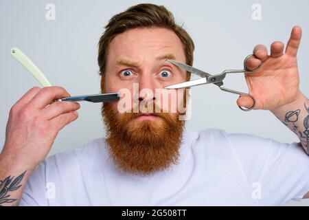 Worried man with scissors and blade is ready to cut the beard Stock Photo