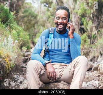 Young African ethnicity man talking via cellphone with somebody and cheerfully laughing as he having a hiking walk in the tropical forest. Happy peopl Stock Photo