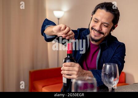 Young happy latin man opening a red wine bottle at home in the living room Stock Photo