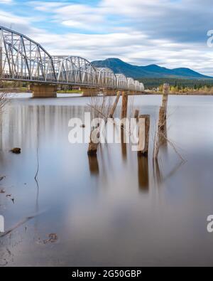 Nisutlin Bay Bridge in Teslin, Yukon Territory, northern Canada with calm water, old wooden posts in the river from the shore. Mountains in background. Stock Photo