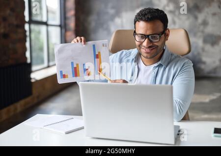 Intelligent indian businessman involved virtual meeting on the laptop sitting in modern office, confident modern businessman in casual wear holding educational course, showing graph to online audience Stock Photo