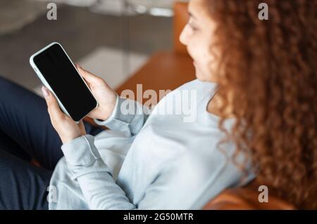 Back view at an empty smartphone screen in hand of a woman, red-haired curly woman holds a mobile phone with blank display, mockup, copy space Stock Photo