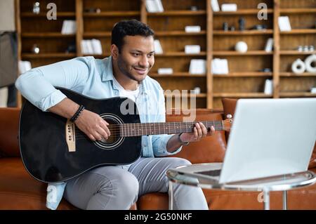 Smiling indian young man learning how to play guitar online, sitting on the sofa in front of the laptop, watching video tutorial, repeats exercises. E-studying and hobby online Stock Photo