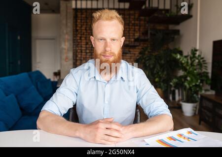 Headshot of redhead businessman in smart casual shirt sis at the desk and looks at the camera with serious face expression, video meeting with red haired guy, portrait of male employee in home office Stock Photo