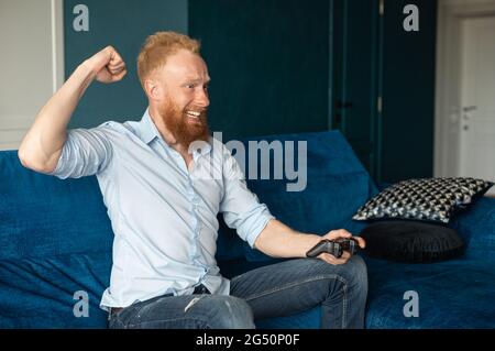 Victory in video game. Overjoyed guy spends leisure time playing on the console. Smiling young redhead man win in video game and feel happy, a guy with a joystick in hand raising fist in triumph Stock Photo