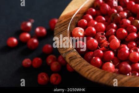 Whole pink red peppercorns in small wooden bowl, some scattered on black gray paper, closeup view from above Stock Photo
