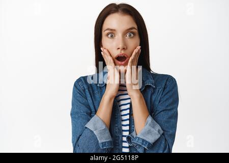 Close up of shocked adult woman gasp, stare startled and speechless at camera, drop jaw and hold hands on face in shock, standing against white Stock Photo
