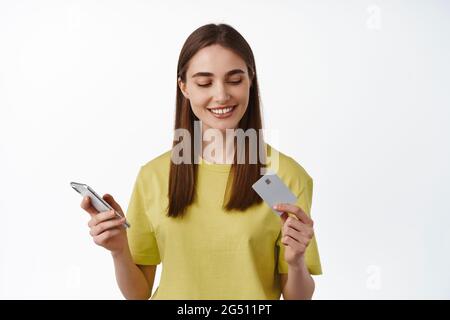 Online shopping. Young brunette woman paying on smartphone, looking at credit card with relaxed smile, purchase on mobile phone,s tanding against Stock Photo