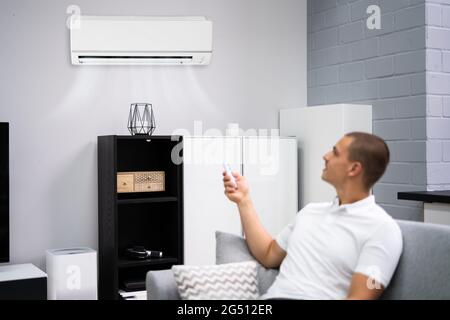 Young Happy Man Sitting On Couch Operating Air Conditioner With Remote Control At Home Stock Photo