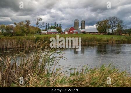 Beautiful farm scene with a red barn and two silos and a pond in the foreground underneath a dramatic sky. Stock Photo