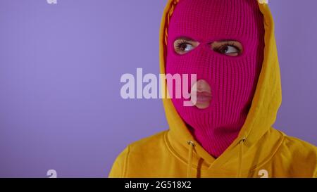 Close up of young woman in pink balaclava and yellow hood pondering about her life on purple background. Thoughtful female in mask looking away Stock Photo