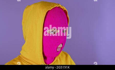Close up of young woman in pink balaclava and yellow hood pondering about her life on purple background. Thoughtful female in mask looking away Stock Photo