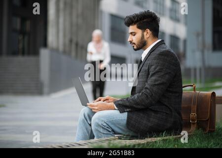 Side view of arabian businessman using modern laptop for remote work while sitting on bench outdoors. Blur background of charming woman in hijab walking and using smartphone. Stock Photo