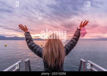 Unrecognized blonde woman in the coast raising her arms and holding her face mask. Horizontal photo Stock Photo