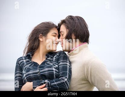 Biracial young couple sitting nose to nose and eyes closed next to ocean outdoors on foggy day Stock Photo