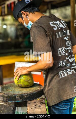 Asian boy wearing a mask opening coconuts with a big knife in a street market in Malaysia Stock Photo