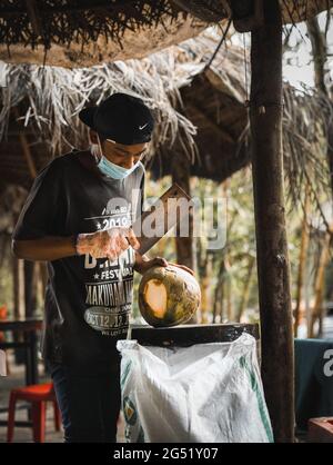 Asian boy wearing a mask opening coconuts with a big knife in a street market in Malaysia Stock Photo
