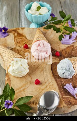 Vanilla, blueberry and raspberry ice cream in waffle cones on wooden board with berries and flowers Stock Photo