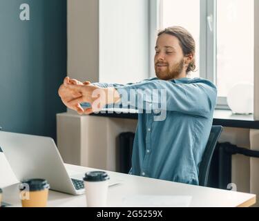 Young tired man office worker in casual clothes stretching arms at workplace Stock Photo