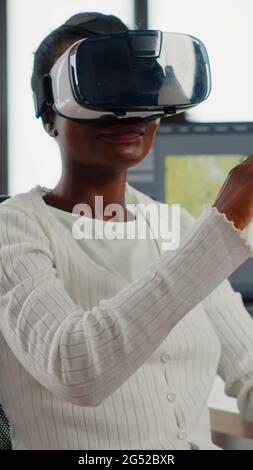Black videographer wearing VR headset editing project in production studio office. Video maker employee processing film montage in digital multimedia company while, colleagues working in background Stock Photo