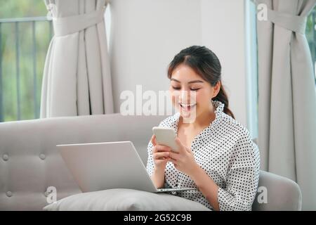 Smiling young asian woman using mobile phone while sitting on a couch at home with laptop computer Stock Photo