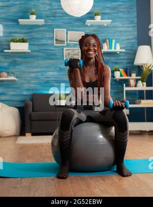 Joyful athletic black person in leggings using stability ball for training biceps, doing curls with dumbbells. Strong athletic person doing sports at home using modern equipment for Stock Photo