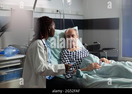 Unwell senior patient laying in bed breathing through oxygen test tube, listening african american doctor specialist, talking about treatment and recovery. Oxymeter attached on finger. Stock Photo