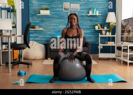 Happy joyful smiling african woman relaxing on swiss ball, after intense hard sport workout on yoga mat in home living room. Cheerful strong athletic fit african using stability ball. Stock Photo