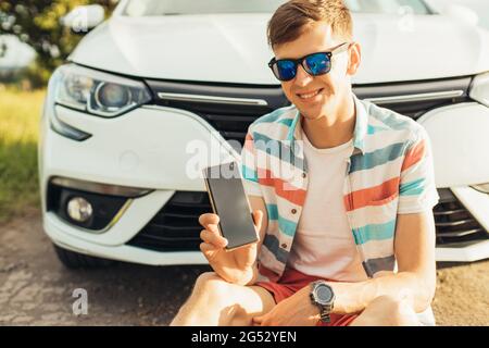 Happy young man in sunglasses showing blank screen of mobile phone while sitting near his car, on summer sunny day Stock Photo