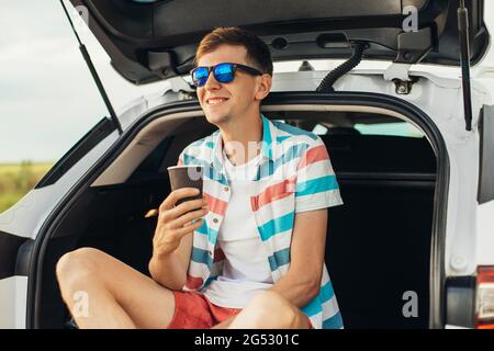 Happy young man, in sunglasses, sits in the trunk of a car, enjoys beautiful nature relaxes and drinks a drink, Summer vacation concept, tourism, trav Stock Photo