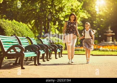 Happy mother and daughter with backpack walking in park on sunny morning. Parent and child going to school together