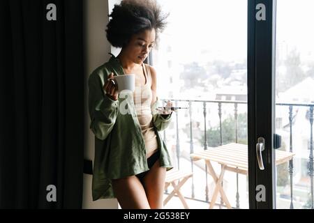 Mid aged african woman holding mobile phone and mug while standing at the window indoors Stock Photo