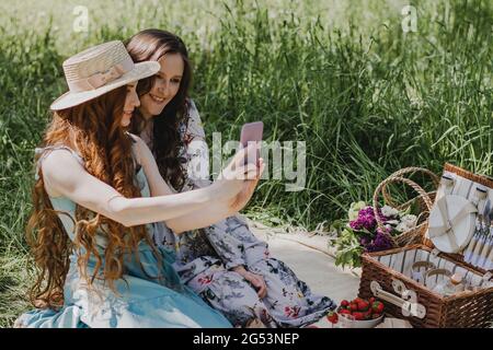 Two beautiful woman in summer dresses enjoying picnic outside and making selfie. Stock Photo